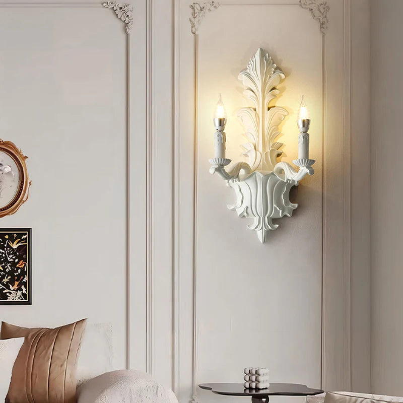 White leaf-shaped wall sconce with two candle-style lights mounted on a paneled wall, near a sofa and a small table.