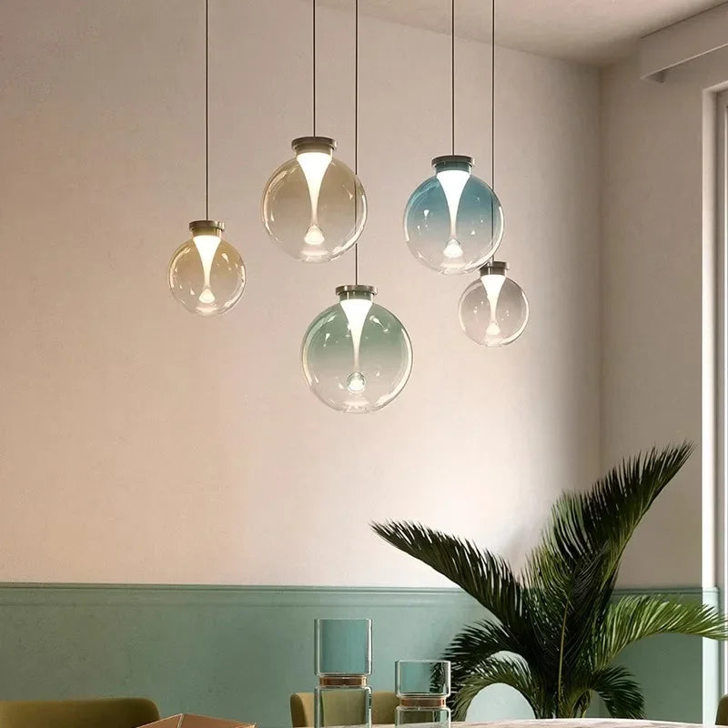 Clear glass globe pendant light with a gradient color wash and a metallic cap, hanging from a black cord against a light wall.