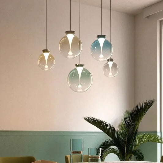 Clear glass globe pendant light with a gradient color wash and a metallic cap, hanging from a black cord against a light wall.