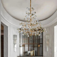 A gold chandelier with crystal floral accents hanging in a well-lit entryway with white curved walls and a large window.