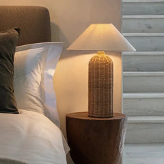 Woven rattan table lamp with a fabric shade placed on a wooden bedside table next to a bed.