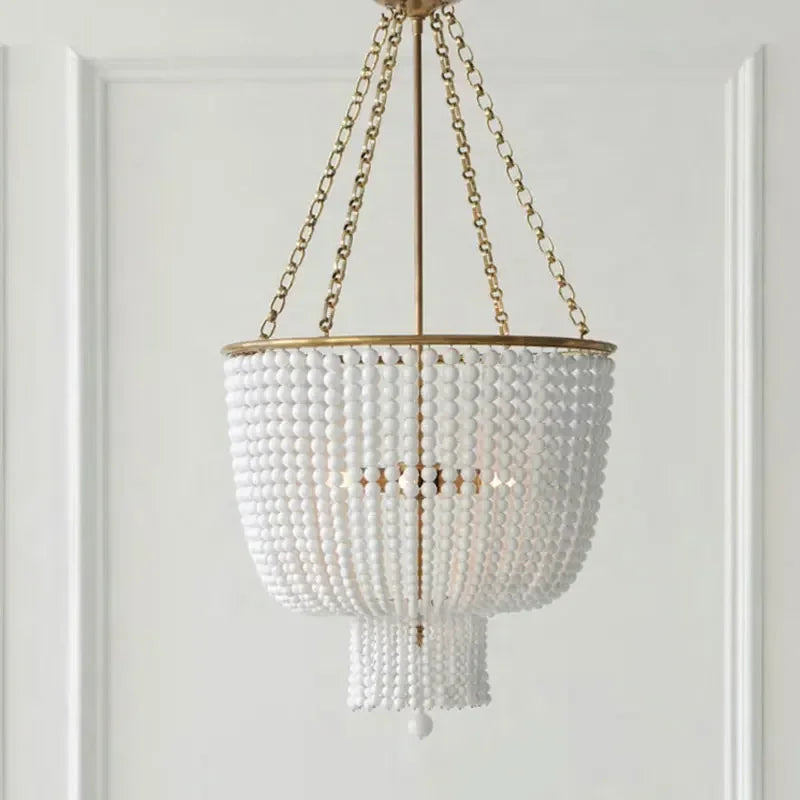 A close-up studio shot of a gold-framed chandelier adorned with numerous white beads, hanging from a gold chain against a white paneled wall.