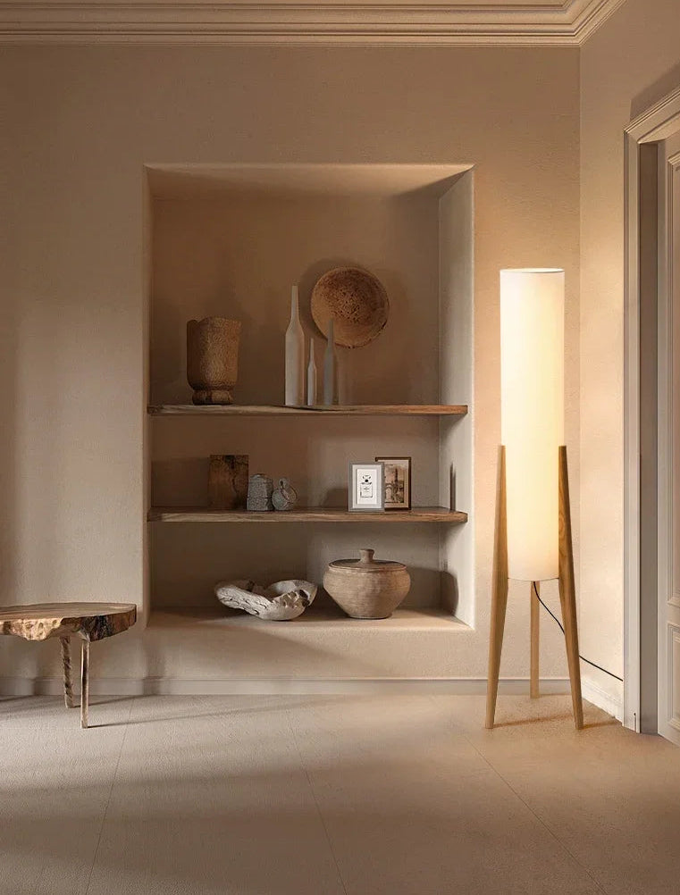 A floor lamp with a black walnut tripod base and a white fabric shade, illuminated in a room with built-in shelves displaying decorative items.