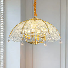 A close-up, front-facing view of a gold-finish chandelier with a fan-shaped crystal-covered canopy and hanging crystal teardrops, illuminated against a softly lit wall.