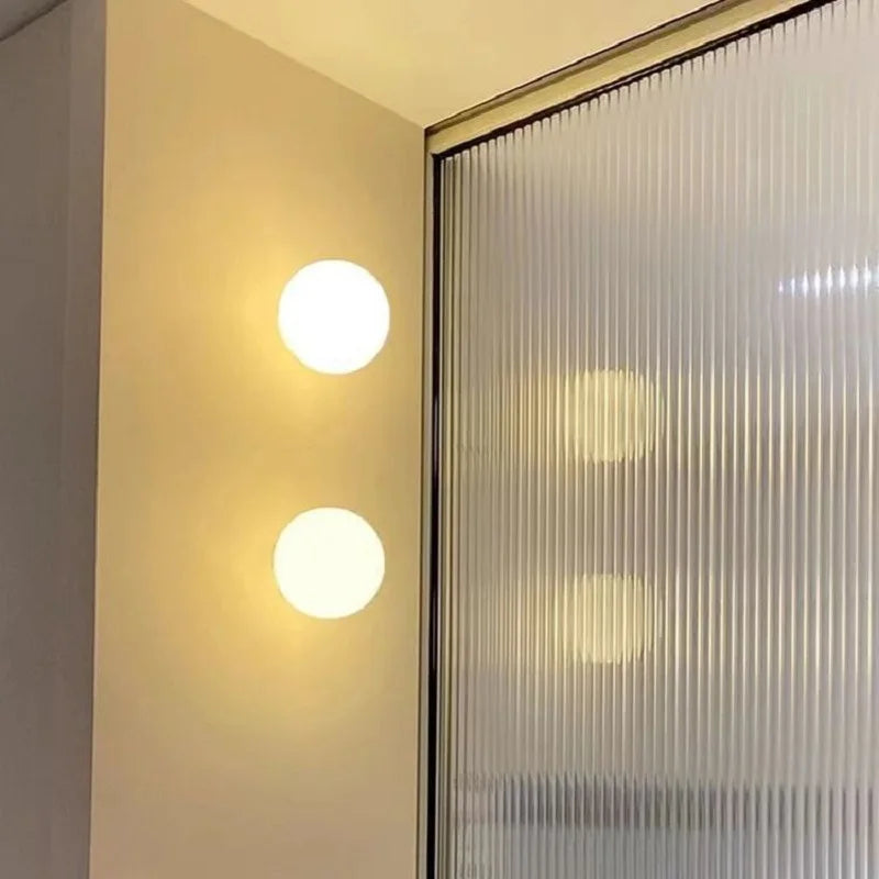 Two round white frosted glass wall lights mounted vertically on a light beige wall next to a ribbed glass partition.