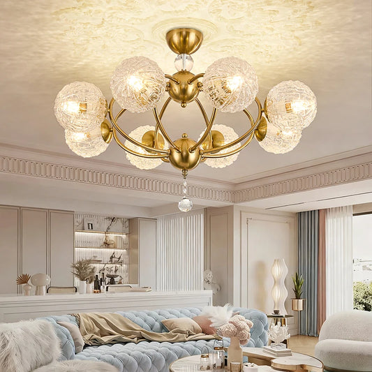 A polished gold chandelier with multiple globe-shaped crystal shades, casting warm light on a white ceiling, installed in a modern living room above a blue sofa.