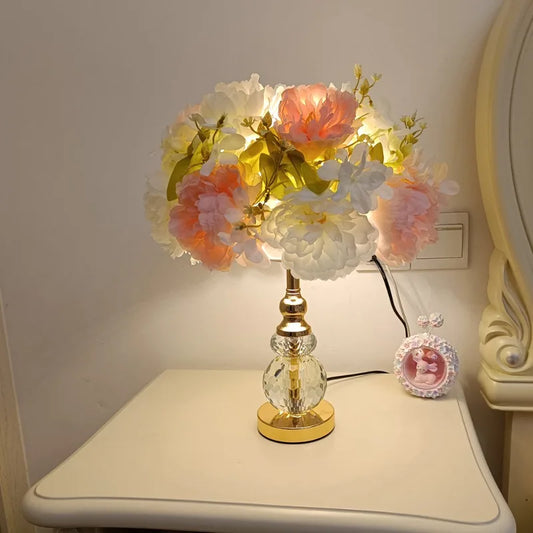 A bedside table with a gold and crystal table lamp featuring a bouquet of pink and white flowers, illuminating the room with warm light.