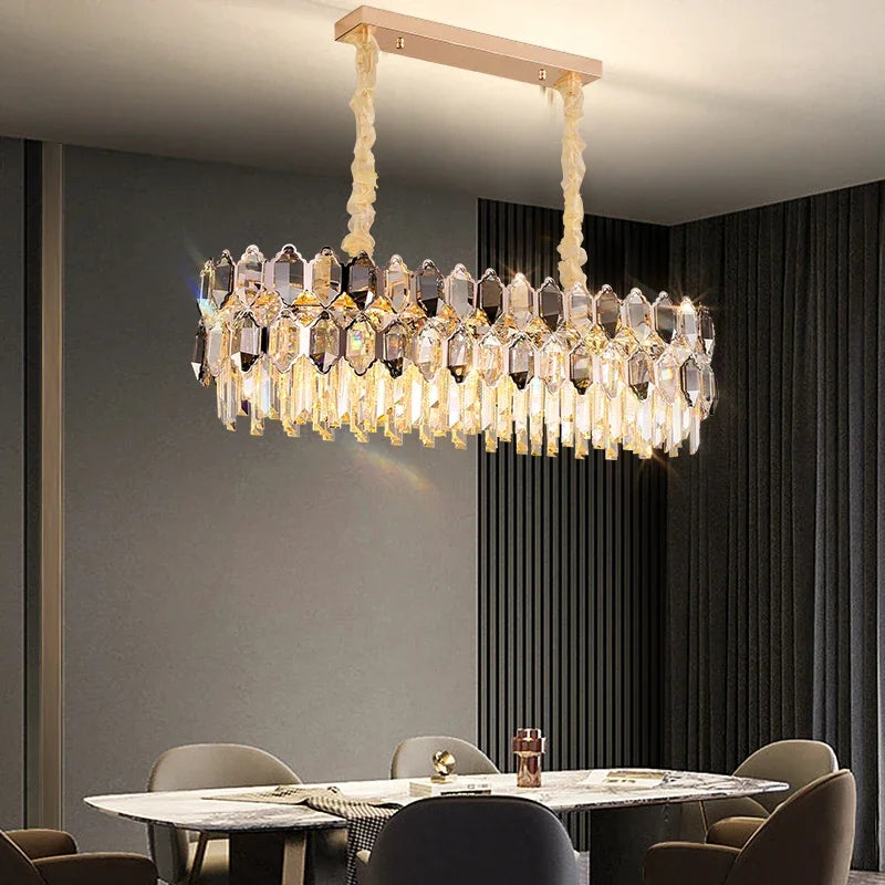 An elongated chandelier with multiple tiers of faceted crystals and a gold-toned frame, hanging over a dining table in a room with dark walls and curtains.
