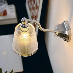 A chrome wall sconce with a textured clear glass shade illuminating a workspace with a laptop and books.
