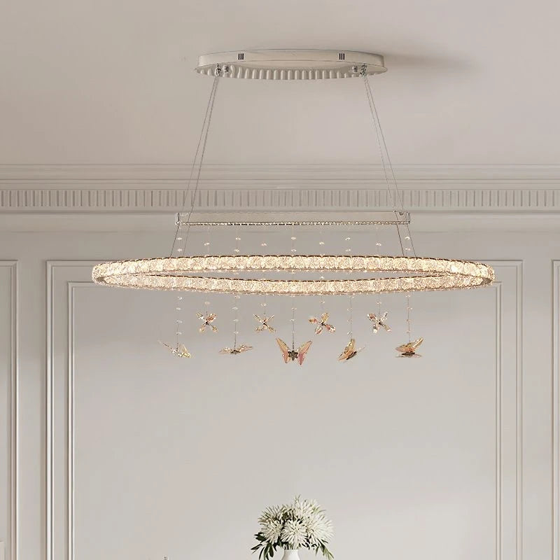 A long, oval-shaped chandelier with multiple rings adorned with clear crystals and hanging butterfly ornaments, suspended from a rectangular ceiling plate.