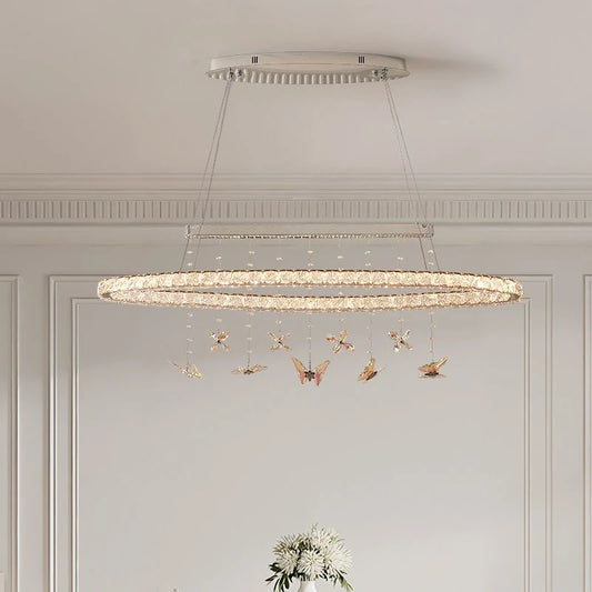 A long, oval-shaped chandelier with multiple rings adorned with clear crystals and hanging butterfly ornaments, suspended from a rectangular ceiling plate.