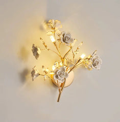 A gold wall sconce with white ceramic flowers and clear crystals, emitting a warm light.