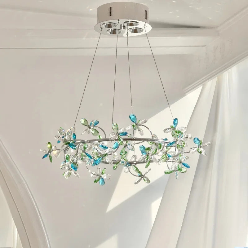 Close-up of a chrome ring chandelier adorned with clear and blue/green crystal flowers, suspended by thin wires against a white ceiling.