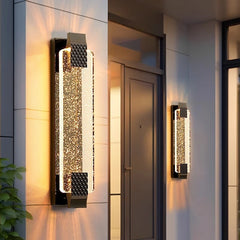 A gold wall sconce with a crystal diffuser illuminated on a marble column next to an entryway.