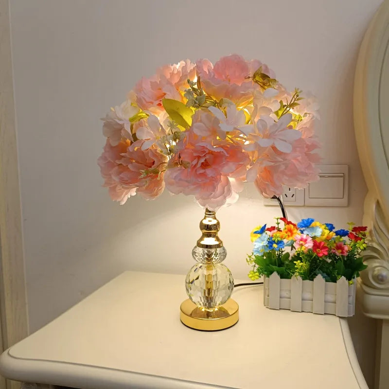 A pink and white floral table lamp with a gold crystal base, placed on a nightstand next to a small potted plant.