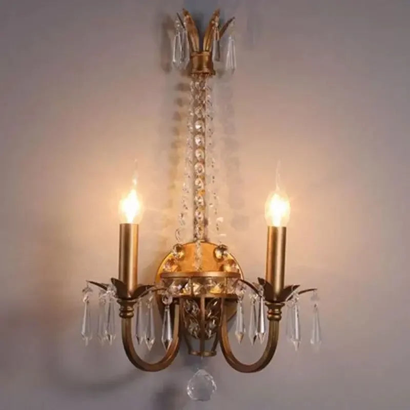 A gold crystal wall sconce with two candle-style lights and ornate detailing, mounted on a gray wall.