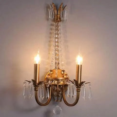 A gold crystal wall sconce with two candle-style lights and ornate detailing, mounted on a gray wall.