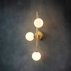 Close-up of a gold wall sconce featuring three round, white frosted glass shades arranged vertically on a brass stem, set against a dark textured wall.