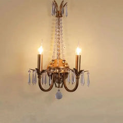 A close-up view of a gold wall sconce with two candle-style lights and dangling clear crystals against a light yellow wall.