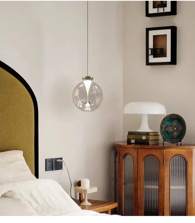 A single clear glass globe pendant light with a metallic cap hanging in a bedroom setting near a bed and a wooden cabinet.