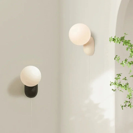 Two minimalist wall sconces with globe shades mounted on a light gray wall, one black and one beige.