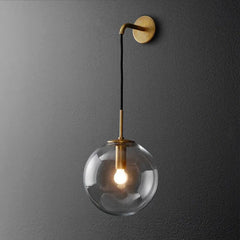 A gold wall sconce with a clear glass globe hanging from an arm mounted on a dark gray wall.