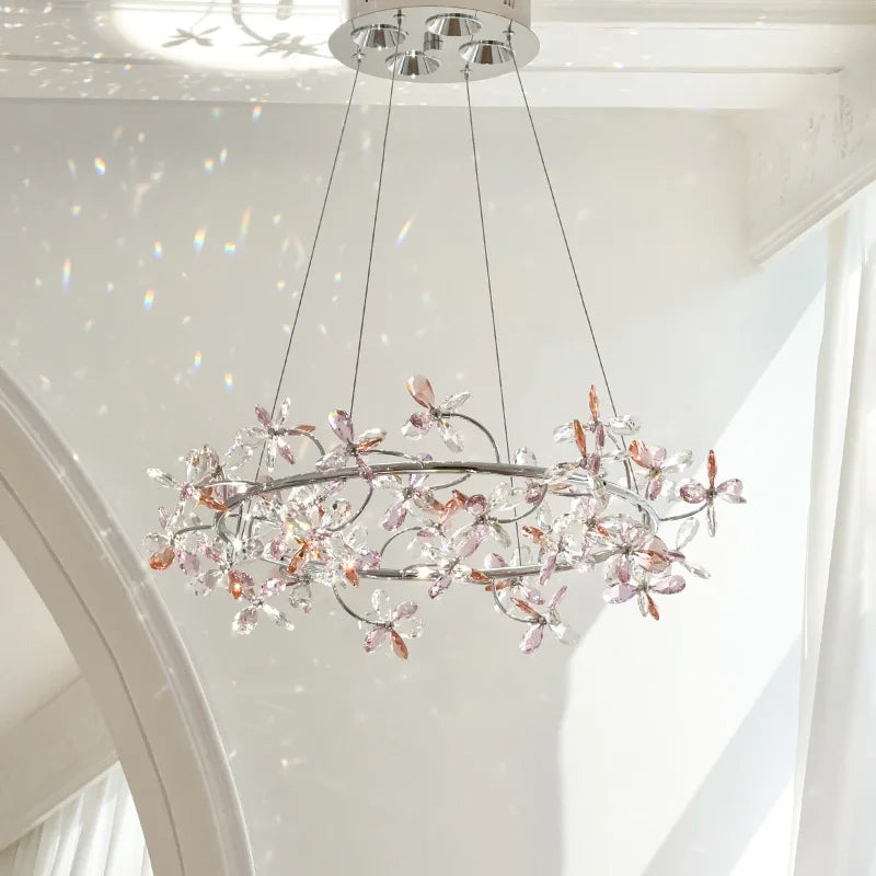 A chrome ring chandelier with crystal floral decorations in varying shades of pink and clear, set against a white ceiling with soft light.