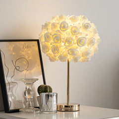 A white floral rose bud table lamp with a gold base and stem, placed on a desk next to a framed picture and a candle.