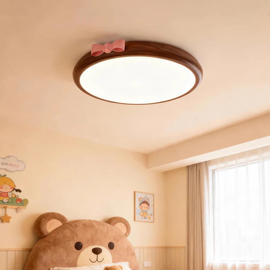 A round, flush mount ceiling light with a wood-tone frame and a pink bow accent, installed in a brightly decorated child's bedroom above a large bear headboard.