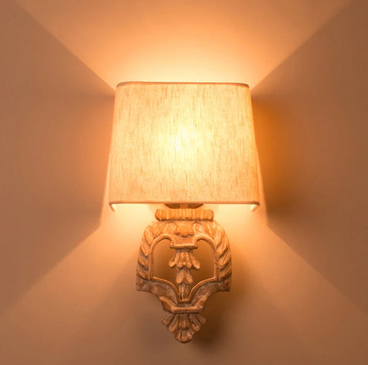 Wall sconce with a carved wooden base and fabric shade illuminated, casting light upwards and downwards on a textured wall.