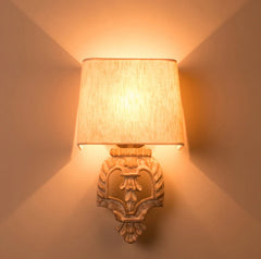 Wall sconce with a carved wooden base and fabric shade illuminated, casting light upwards and downwards on a textured wall.