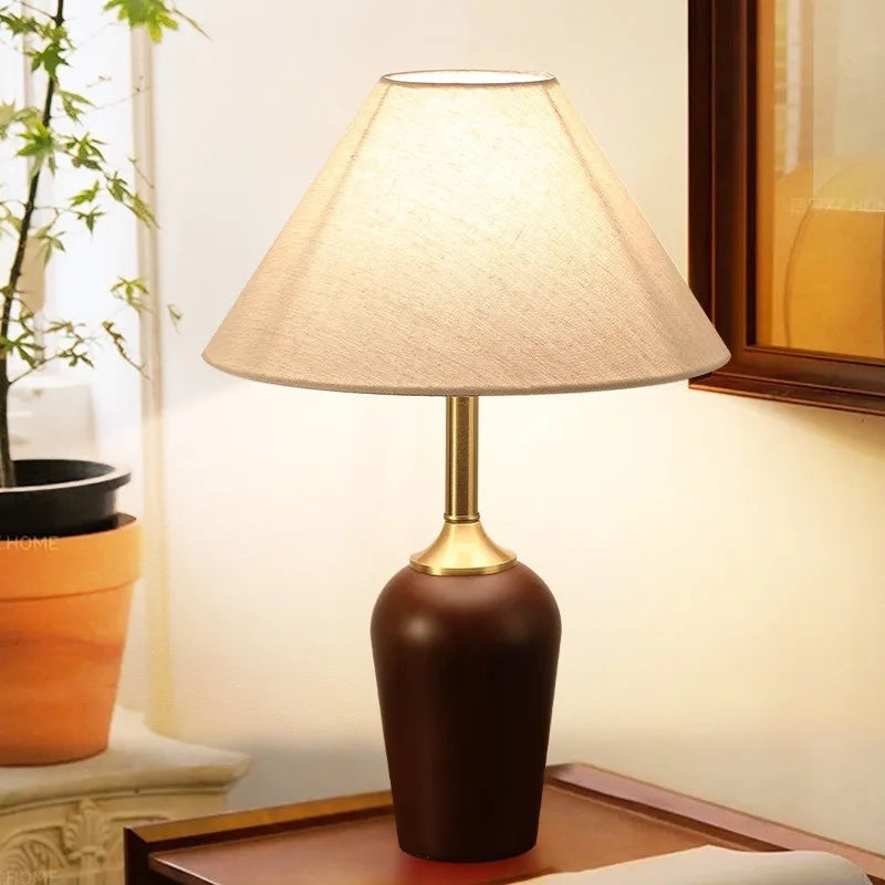 Table lamp with a brown base and beige shade on a wooden surface, next to a plant.