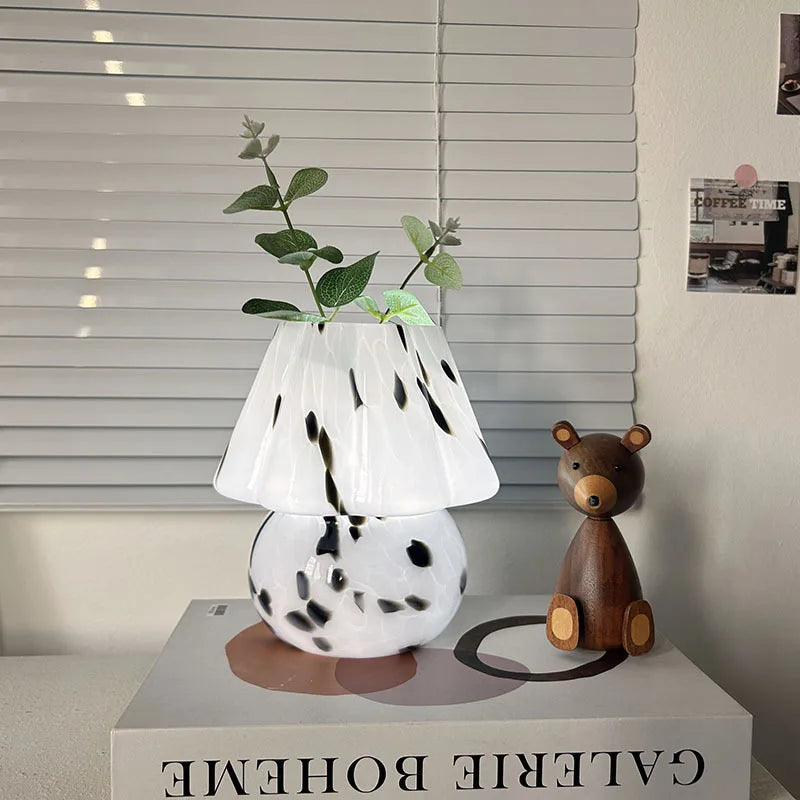 White lamp with black spots on a box next to a wooden bear toy, with a blurred window and poster in the background.