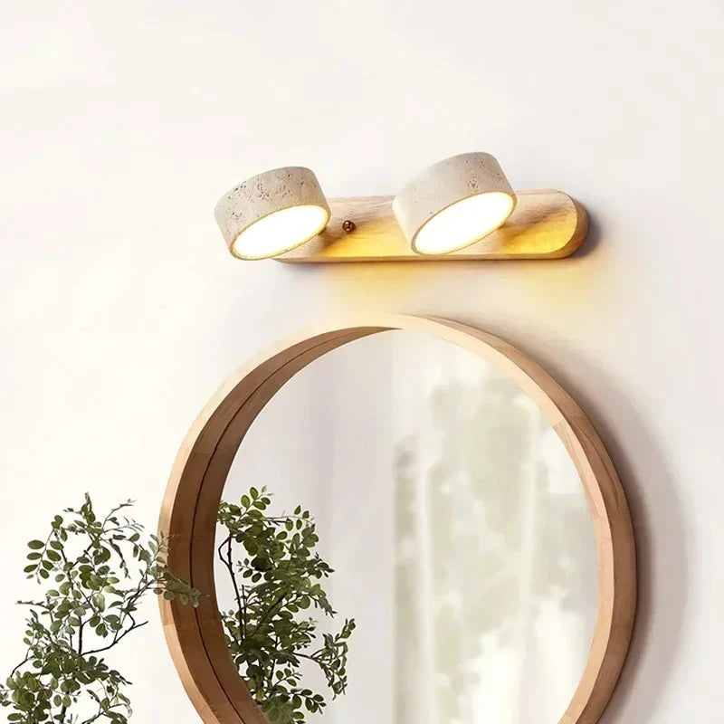 Close-up of a wooden base wall sconce with two stone cylinder shades, positioned above a round mirror with greenery in the foreground.