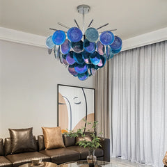A modern chandelier with layered blue glass discs suspended from a chrome frame, casting light in a room with a brown leather sofa.