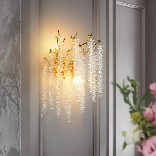 A close-up view of a gold crystal branch wall sconce mounted on a wall, with cascading clear crystals illuminated by warm light.