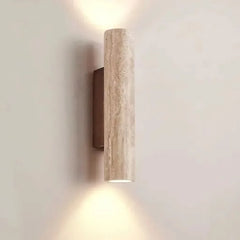 A single cylindrical wall sconce made of textured, light-colored stone, mounted on a dark bracket, emitting light upwards and downwards against a plain wall.