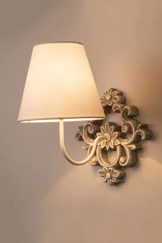 Close-up of a gray-white wooden wall sconce with a fabric shade and decorative floral accents against a muted background.