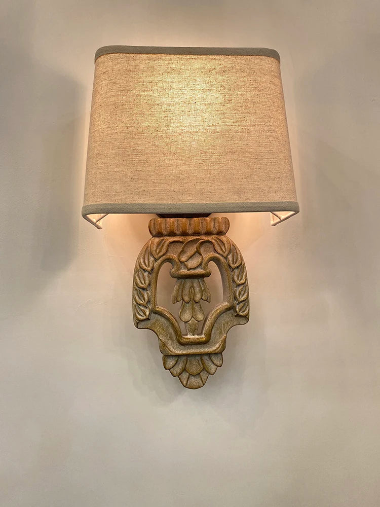 Close-up of a vintage-style wall sconce with a carved wooden base and a light beige fabric shade mounted on a white wall.