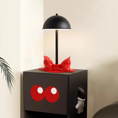 Black dome shade table lamp with a red bow accent on a dark cabinet with large red eyes.