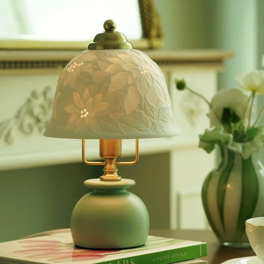 Table lamp with a white ceramic dome shade featuring embossed floral patterns and a green base, positioned on a book with a vase of flowers in the background.