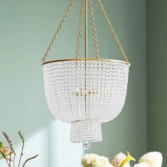 A clear crystal chandelier with a gold frame is displayed in a room with pale green walls and a table with flowers in the foreground.