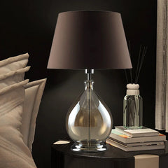 Amber glass table lamp with dark fabric shade on a nightstand next to a bed, with books and a diffuser in the background.