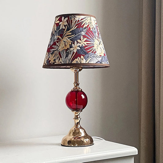 A polished brass table lamp with a conical floral fabric shade and a round red glass orb accent, lit and placed on a white surface against a neutral backdrop.