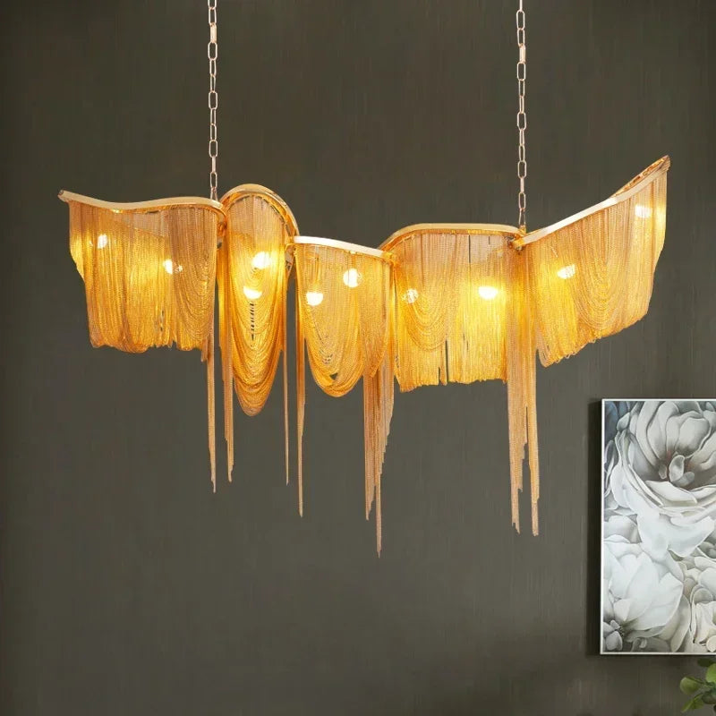 Gold aluminum chain tassel pendant light with cascading elements, suspended by chains against a dark wall.
