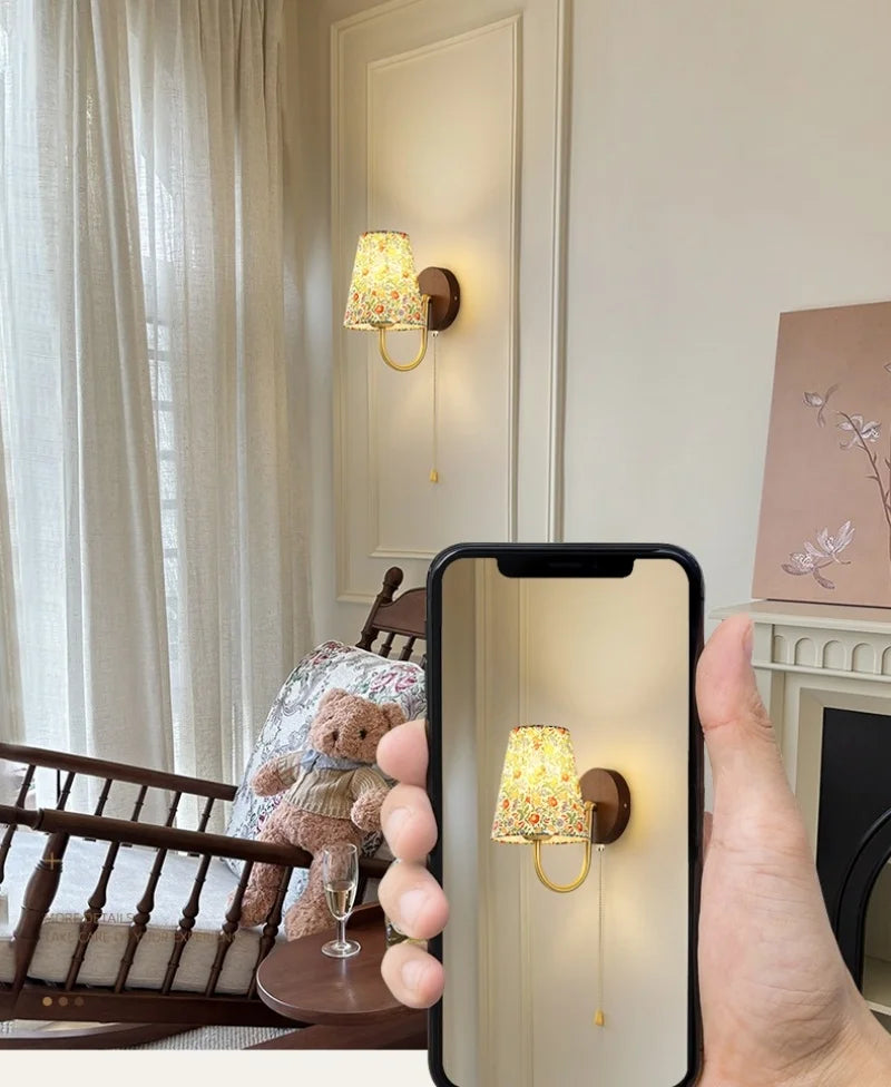 Hand holding a phone showing a wall-mounted lamp design in a room with a teddy bear and chair.