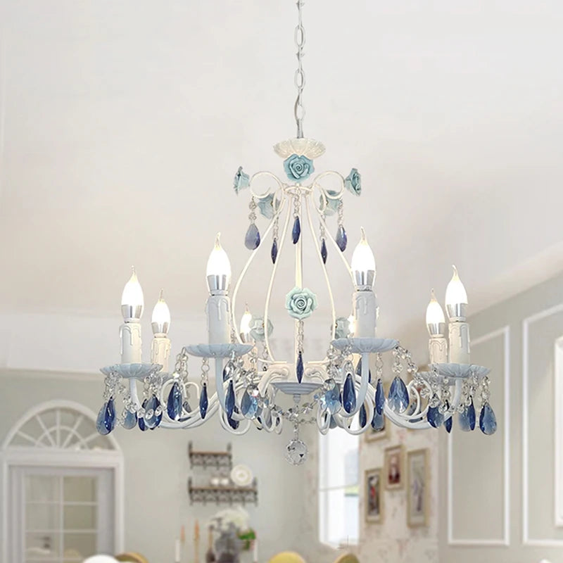 White metal chandelier with blue floral accents and blue crystal drops, illuminated and hanging from a white ceiling in a dining room setting.