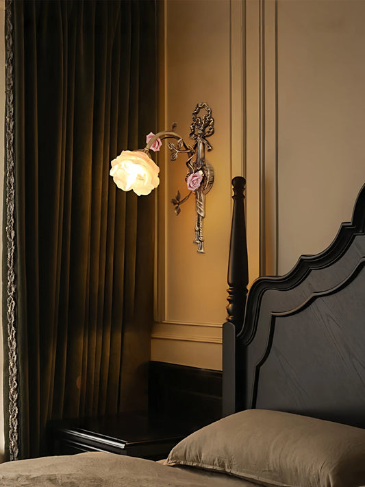Antique gold flower wall sconce with a white frosted glass shade, casting a warm glow in a dimly lit bedroom next to dark green curtains and a dark wood bed.