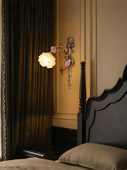 Antique gold flower wall sconce with a white frosted glass shade, casting a warm glow in a dimly lit bedroom next to dark green curtains and a dark wood bed.