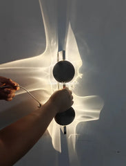 Hands adjusting a wall sconce with two black discs, highlighting its mounting and lighting effect.